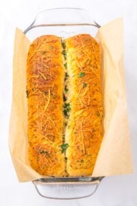 Two golden brown loaves of herb and cheese bread in a parchment-lined glass dish.