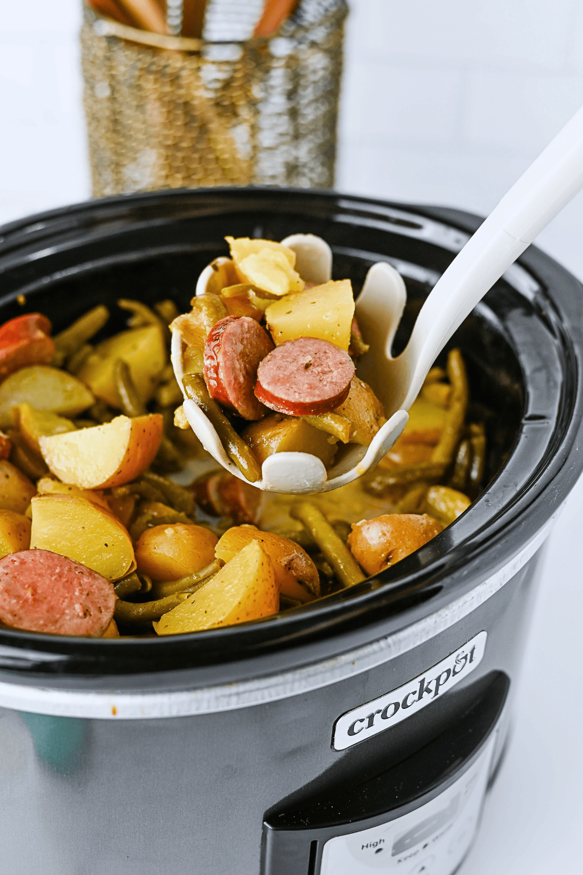 A ladle serving sausage, potatoes, and green beans from a crockpot.