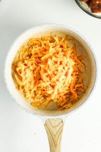 Shredded cheddar cheese in a white saucepan on a white surface.