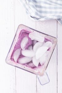 Top view of a blender with a purple smoothie and ice cubes on a white table.