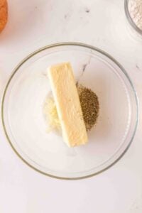 A glass bowl with a stick of butter and dried herbs on a white surface.