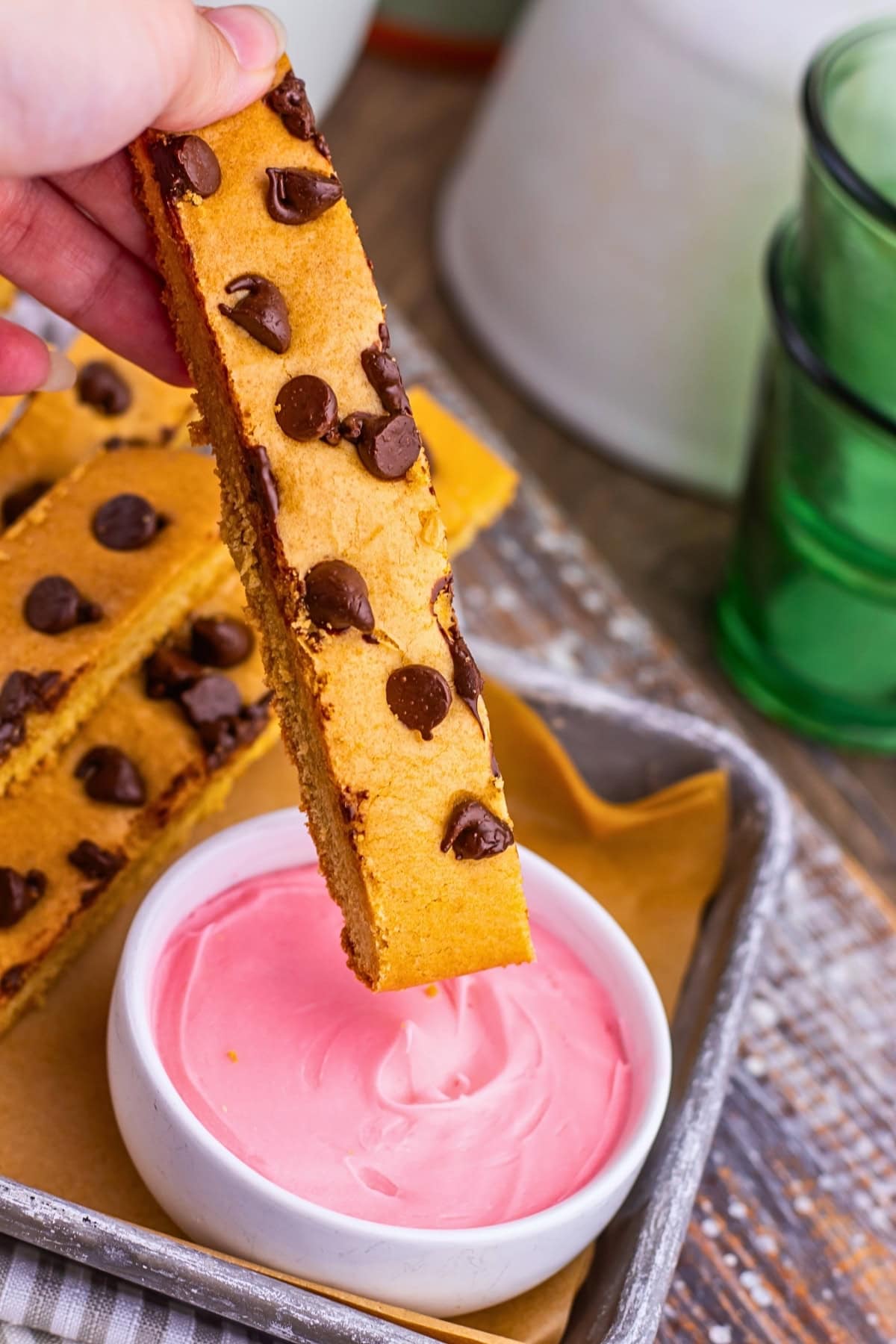 A hand dips a chocolate chip cookie stick into a bowl of pink frosting.