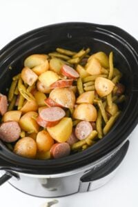 Sliced potatoes, green beans, and sausage cooked in a black slow cooker.