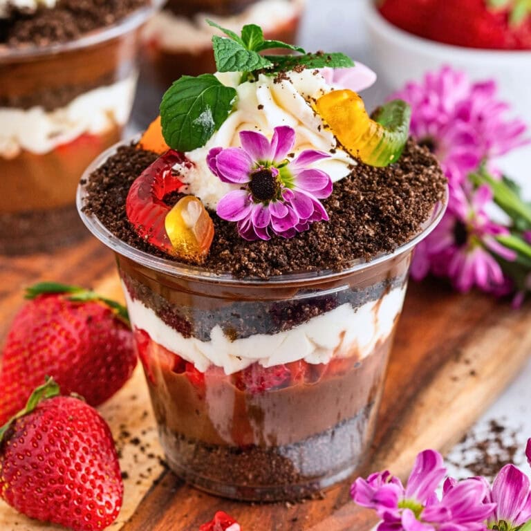 Chocolate dessert cup with cream, gummy worms, flowers, and strawberries beside it.