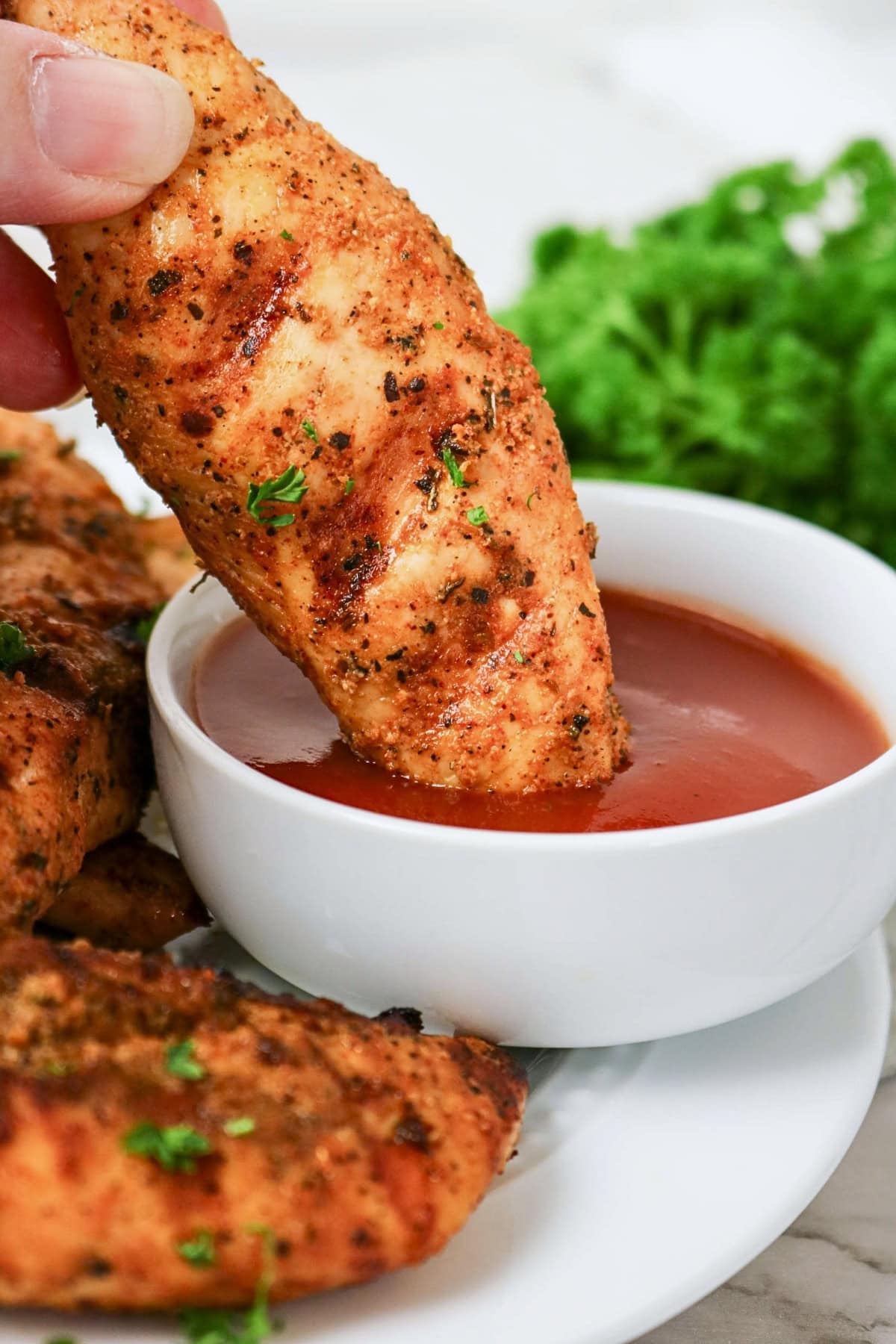 Grilled chicken tender being dipped into red sauce, with parsley in background.