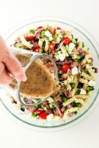 Dressing being poured over a bowl of pasta salad with veggies and cheese.