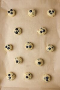 Scoops of cookie dough with blueberries on a parchment-lined baking sheet.