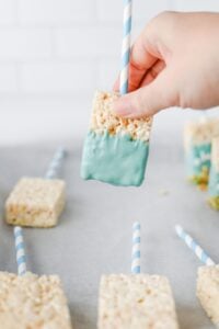 A hand holds a rice krispie treat on a stick dipped in blue coating.