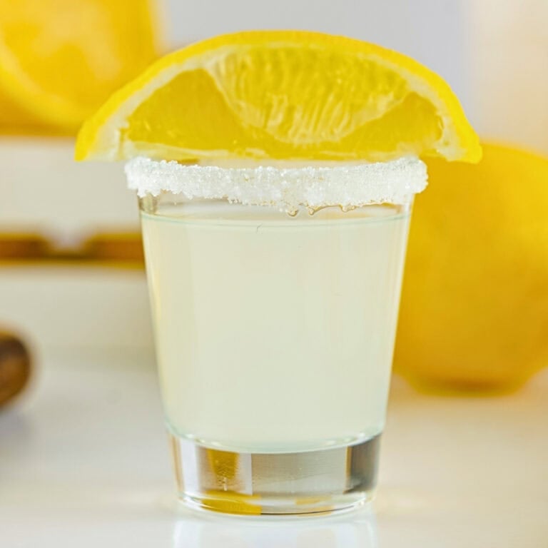 A shot glass with a sugar rim, lemon drink, and a lemon wedge garnish.