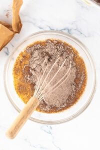 A whisk in a bowl mixing brown dry ingredients with wet ingredients on a marble surface.