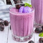A glass of blackberry smoothie with purple ice cream, berries, and mint on top.