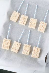 Eight Rice Krispies treats with blue striped sticks on parchment paper.