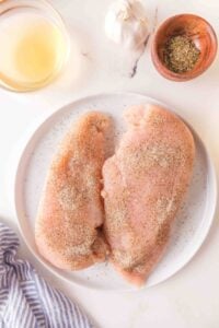 Raw seasoned chicken breasts on a white plate with herbs, garlic, and wine nearby.