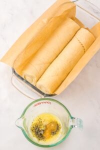Two rolls of dough in a parchment-lined dish, with melted butter and herbs in a cup.