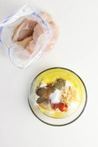 Raw chicken in a plastic bag next to a bowl of mixed spices and yogurt marinade.