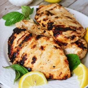 Grilled chicken breasts on a plate with lemon slices and fresh basil leaves.