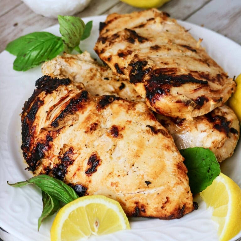 Grilled chicken breasts on a plate with lemon slices and fresh basil leaves.
