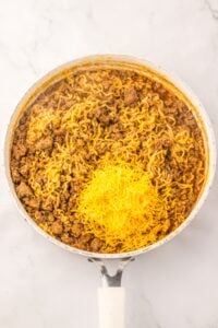 A skillet with cooked ramen, ground beef, and a pile of shredded cheddar cheese.