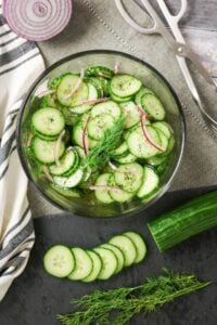 Glass bowl of cucumber salad with red onions and dill, surrounded by fresh ingredients.