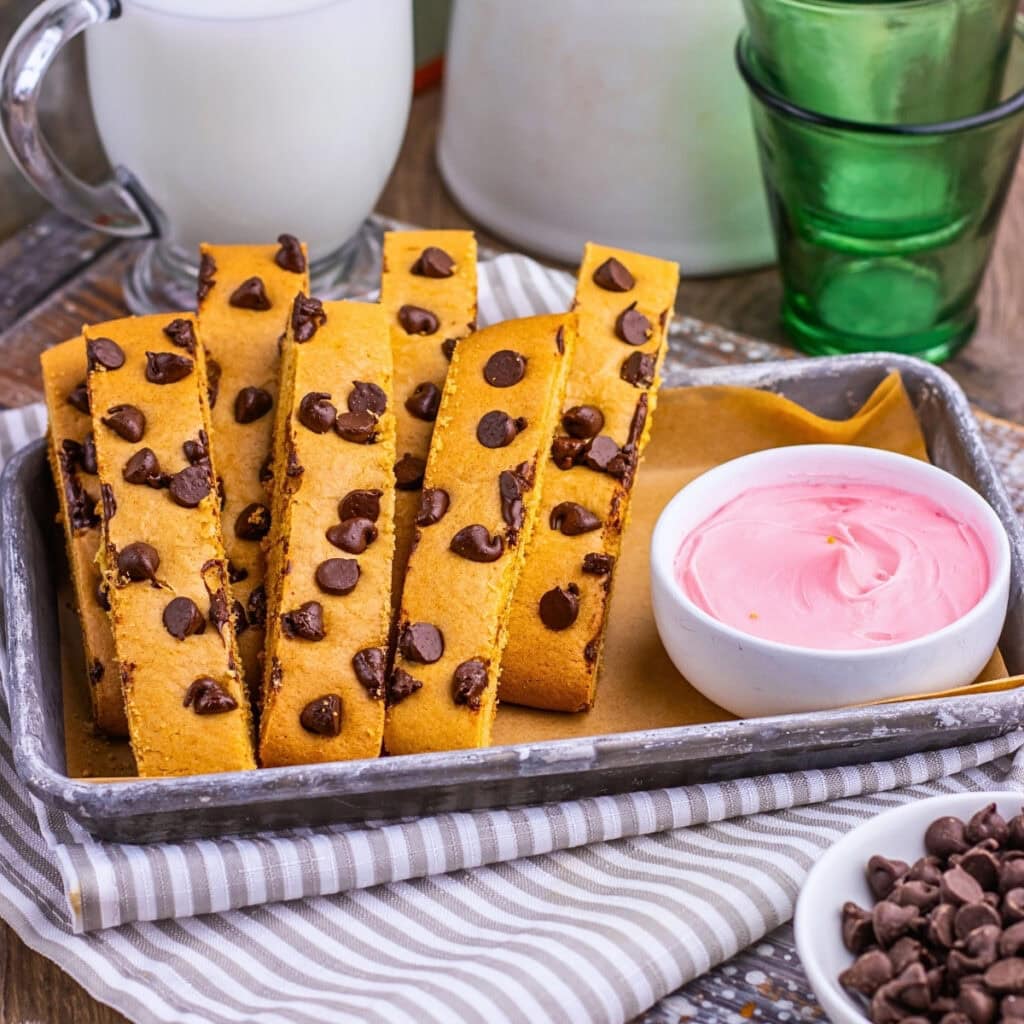 Chocolate chip cookie sticks on a tray with pink dip, milk, and green glasses.