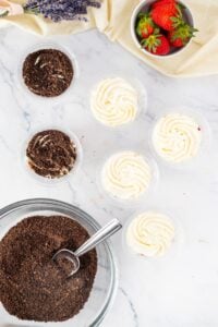 Six dessert cups with cream, crushed cookies, strawberries, and a mixing bowl.