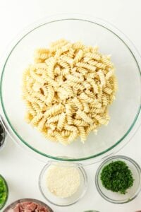 Glass bowl of uncooked rotini pasta surrounded by small bowls of ingredients.