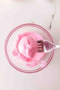 A fork holds a round treat dipped in pink coating over a glass bowl.