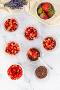 Cups of chocolate pudding topped with chopped strawberries on a marble surface.