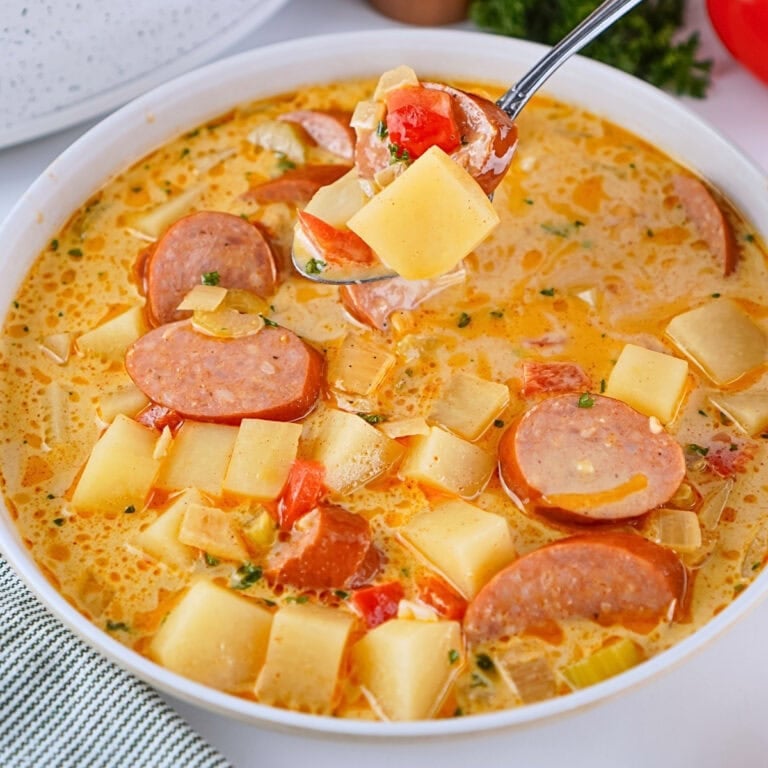 A bowl of creamy sausage and potato soup with vegetables, held by a metal spoon.
