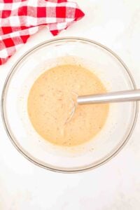 Glass bowl of batter with a whisk, next to a red and white checkered cloth.