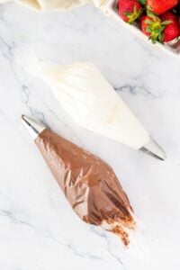 Two piping bags with cream and chocolate frosting, next to fresh strawberries.