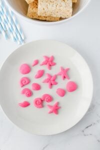 Pink candy sea shells and starfish on a white plate, straws and treats nearby.