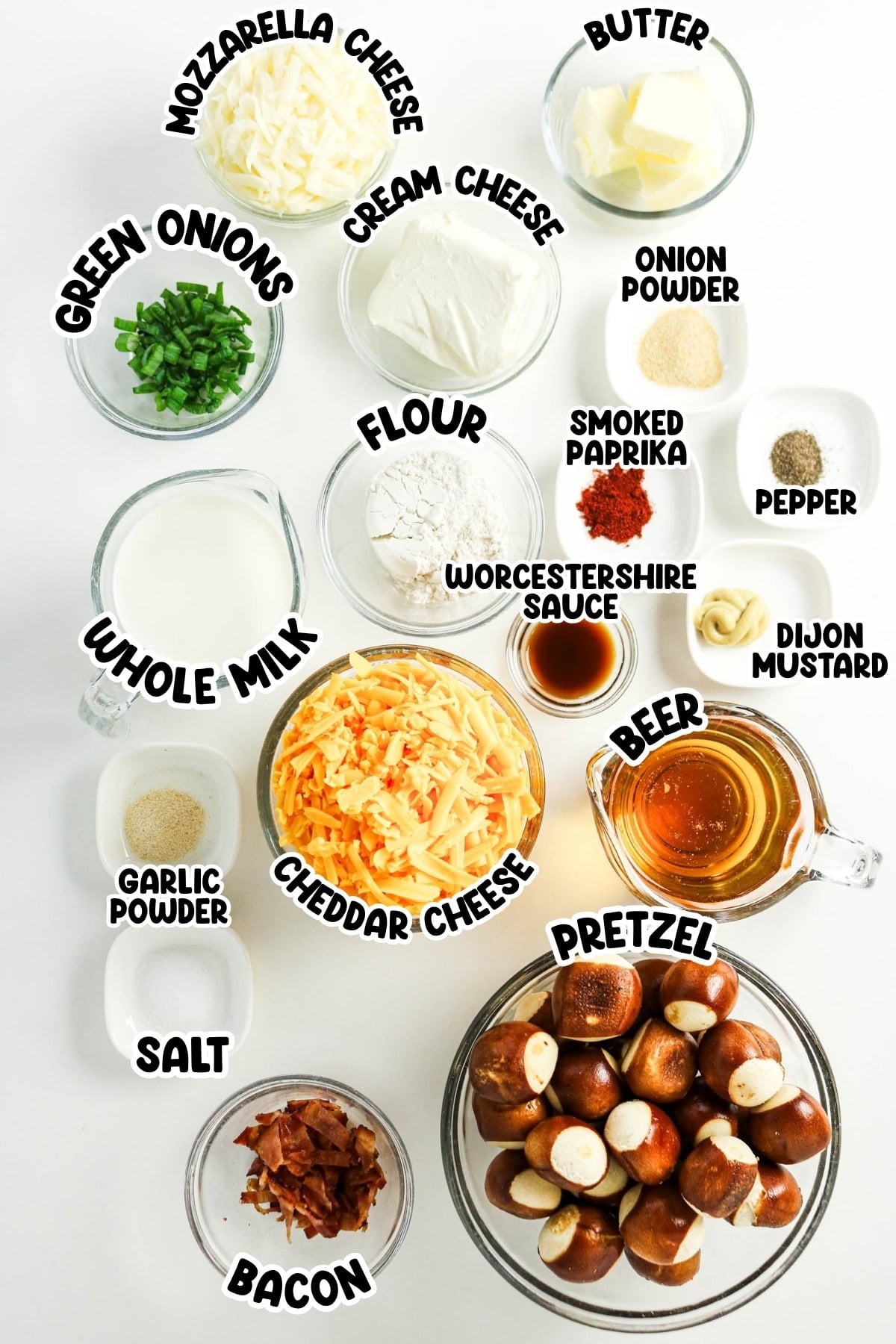 Assorted ingredients for cheese dip and pretzels arranged in glass bowls on a white table.
