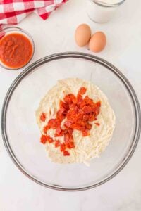 A glass bowl with flour, cheese, and chopped pepperoni; eggs and sauce nearby.