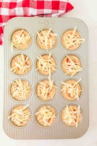 A muffin tin filled with batter and shredded cheese, ready to be baked.