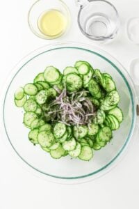 Sliced cucumbers, red onions, and herbs in a glass bowl with dressing nearby.