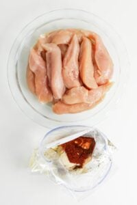 Raw chicken strips on a plate and spices in a plastic bag on a white surface.