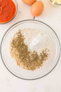 A glass bowl with flour, herbs, and baking powder, with eggs and sauce nearby.