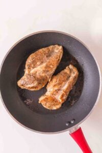 Two seasoned chicken breasts cooking in a black frying pan with a red handle.
