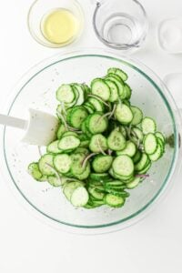 Sliced cucumbers and onions in a glass bowl with dressing ingredients nearby.