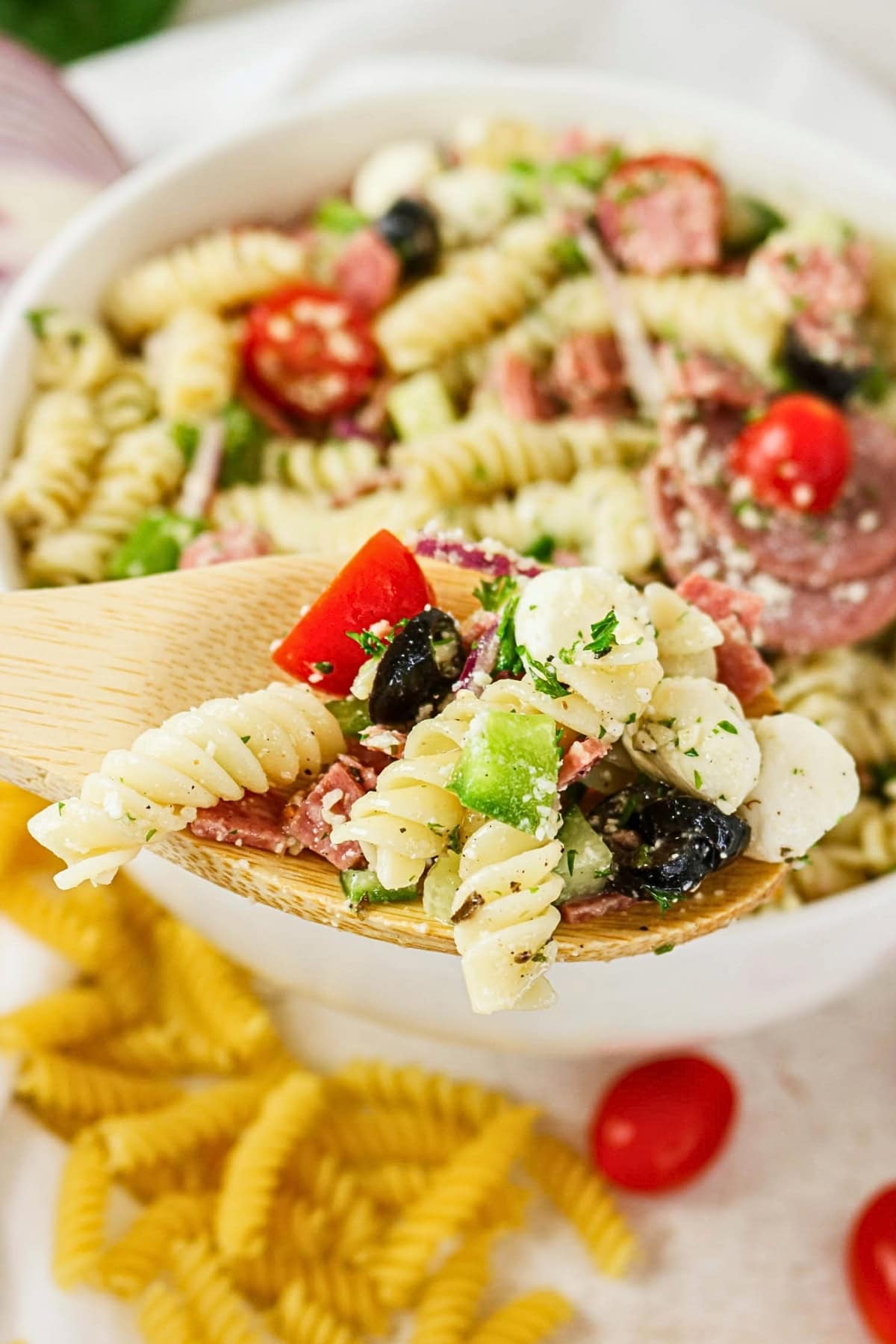 A wooden spoon holds pasta salad with veggies, cheese, and olives above a bowl.