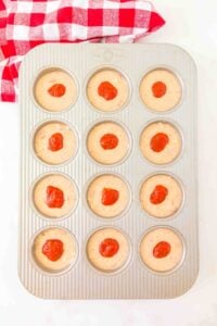 Muffin tin with batter in cups, each topped with a dollop of red sauce.