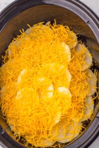 Sliced potatoes topped with shredded cheddar cheese in a slow cooker.