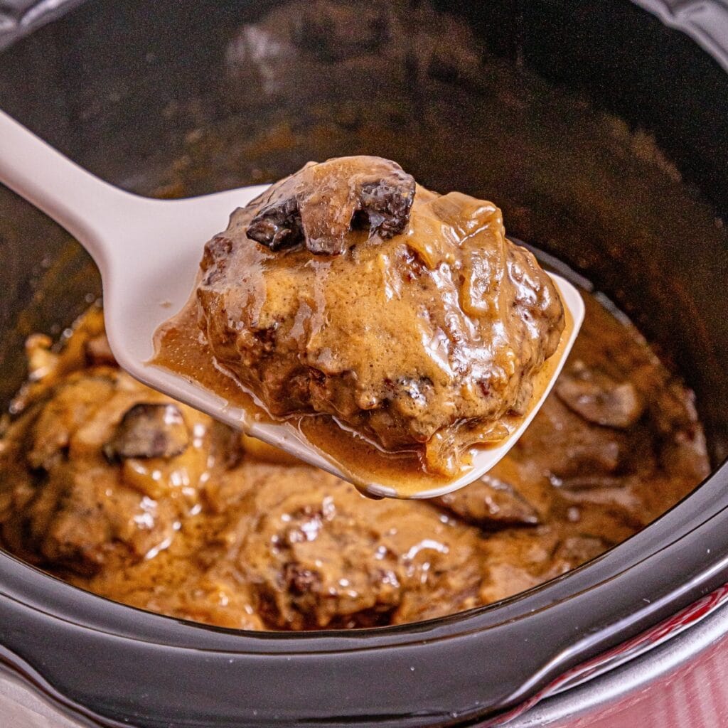 A spoon lifts saucy Salisbury steak with mushrooms from a slow cooker.