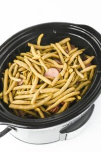 Fresh green beans and sausage slices in a black slow cooker on a white background.