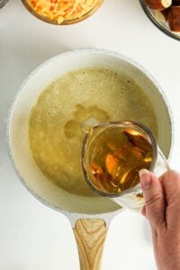 A hand pours beer from a measuring cup into a white saucepan on a stove.