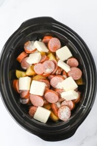 Sliced sausage, potatoes, and butter in a black slow cooker, ready to cook.