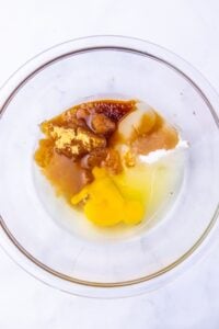 Clear bowl with eggs, sugar, brown sugar, vanilla, and butter on a white background.