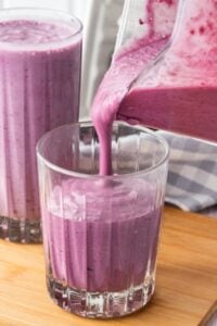 A purple smoothie being poured into a glass on a wooden surface.