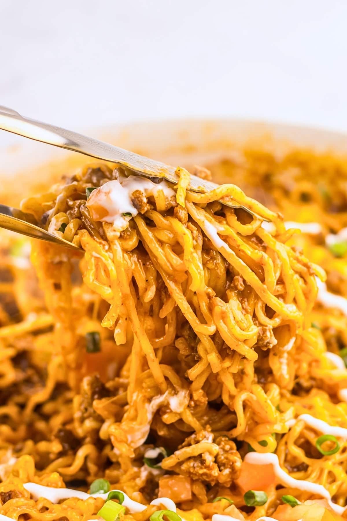 Tongs lifting cheesy ramen noodles with ground meat and chopped green onions.